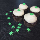 Four chocolate cupcakes with white frosting, each topped with a small green leaf decoration made from The Spice Lab Hemp Leaf Sprinkles CBD & THC Free by The Spice Lab, are arranged on a dark surface scattered with more edible party decorations.