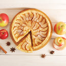 A round apple tart with a slice cut out sits on a wooden board, surrounded by fresh apples, cinnamon sticks, star anise, and an apple peel on a light wooden surface—a perfect scene to showcase the warmth of The Spice Lab Apple Pie Spice Blend and the inviting aroma of seasonal spice from The Spice Lab.