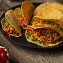 Three tacos filled with ground beef seasoned with The Spice Lab Organic Chili Powder, shredded cheese, lettuce, and chopped tomatoes on a black plate, set on a rustic wooden table.
