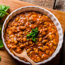 A round white dish filled with baked beans in a rich sauce, enhanced with a hint of The Spice Lab Ancho Chile Powder by The Spice Lab and garnished with chopped green onions, sits on a wooden surface alongside a small wooden spoon and some spinach leaves.