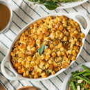 A white oval dish filled with golden, crispy bread stuffing—expertly seasoned with The Spice Lab Organic Rubbed Sage—garnished with a sage leaf, placed on a striped cloth, with green beans and gravy nearby.