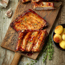 Barbecued ribs with herbs on a wooden cutting board, surrounded by The Spice Lab Organic Minced Garlic, peppercorns, and a bowl of potatoes, all set on a rustic wooden table.
