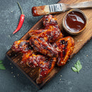 Grilled chicken wings glazed with barbecue sauce on a wooden board, served with a bowl of sauce, a brush, a red chili pepper, and parsley leaves, garnished with sesame seeds and brought to life by The Spice Lab Organic Granulated Onion—a pantry staple from The Spice Lab that adds depth and flavor to every bite.