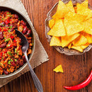 A bowl of chili with beans and ground meat, seasoned with The Spice Lab Organic Chili Powder Blend, sits beside a plate of yellow tortilla chips on a wooden table, with a red chili pepper and a single chip nearby.