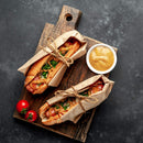 Two hot dogs wrapped in brown paper and tied with twine rest on a wooden board next to a small bowl of The Spice Lab Organic Ground Mustard and two cherry tomatoes, all arranged on a dark surface. This classic presentation is perfect for pairing with your favorite spice blends from The Spice Lab or serving alongside cheese dishes.
