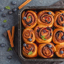 A baking tray of blueberry cinnamon rolls garnished with fresh blueberries and mint leaves, baked to perfection with The Spice Lab Organic Ground Vietnamese Saigon Cinnamon for a rich, aromatic flavor; cinnamon sticks and loose blueberries are artfully arranged on a dark surface nearby, highlighting the premium quality of The Spice Lab product.
