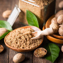 A rustic wooden surface displays The Spice Lab Organic Ground Nutmeg in a wooden bowl, accompanied by a small wooden scoop, whole nutmeg seeds, fresh green leaves, a grater, and a glass jar—perfect for infusing your holiday baking or classic eggnog with warm, aromatic flavor from The Spice Lab.