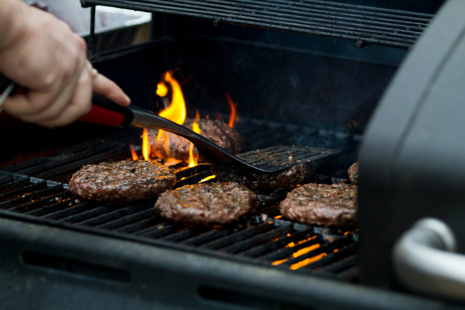 Six beef patties cooking on a grill with flames rising beneath them. A hand uses a spatula to flip one of the patties. The grill lid is partially open.