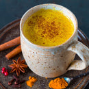 A mug filled with The Spice Lab Golden Milk Turmeric Tea Superfood Premium Powder sits on a plate, accompanied by cinnamon sticks, star anise, cloves, red berries, and a spoonful of turmeric—a cozy scene that highlights the nourishing warmth of this ancient superfood from The Spice Lab.