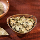 A wooden bowl brimming with The Spice Lab Cracked Bay Leaves 1 lb Bulk Foodservice Herb, ideal for Mediterranean cuisine, rests on a wooden surface, accompanied by part of another bowl and a loose bay leaf from The Spice Lab nearby.