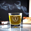 A short, square-bottomed Whiskey Glasses: Modern by Swanky Badger filled with amber liquid and etched with an S-logo inside a geometric design, surrounded by wisps of smoke.