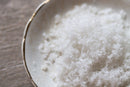 A close-up of World Spice Fleur de Sel sea salt flakes from World Spice, piled on a white ceramic plate with a gold rim, set on a wooden surface.