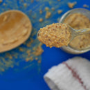 A close-up of a spoonful of SALTOPIA French Mustard Salt: MUSTARD BEEN LOVE held above its jar, with Dijon mustard salt smears, a wooden spoon, and a white cloth with a red stripe on a blue surface in the background.