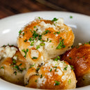A close-up of golden brown garlic knots topped with SALTOPIA Infused Sea Salts’ Garlic Salt: MOUTH FULL OF MAGIC and chopped parsley, served in a white bowl.