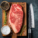 The Dude Food Gentleman’s Meat Box by Dude Food features raw marbled Kansas City Strip Steak displayed on a wooden cutting board, accompanied by coarse salt, peppercorns, rosemary, thyme, and a large chef’s knife on a dark countertop.