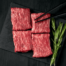 Four slices of marbled raw beef from the Dude Food Gentleman’s Meat Box by Dude Food are arranged on a dark slate board with a sprig of rosemary beside them. Black chopsticks rest on one slice, all set against a black wooden background.