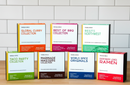 Seven World Spice Best of the Northwest collection boxes from World Spice are arranged in two rows on a wooden surface with a white tiled background. Each box showcases a unique theme—including curry, BBQ, ramen, and taco party—and features flavors inspired by classic Pacific Northwest spices such as smoked salt and Seattle seasoning.