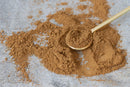 A wooden spoon rests on a gray surface dusted with scattered brown World Spice Gingerbread Spice from World Spice, evoking the warm aroma of gingerbread and inviting thoughts of spicy baking.