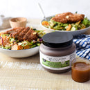 A quart bottle of Terrapin Ridge Farms Ginger, Miso & Honey Dressing sits on a wooden table beside a small glass of dressing and two salad bowls with breaded chicken and orange slices. A blue checkered napkin is nearby.