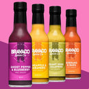Four bottles of Bravado Spice Co hot sauce with vibrant labels—Ghost Pepper & Blueberry, Pineapple & Habanero, Creamy Herb Jalapeno, and Serrano & Basil—are displayed against a pink background. Featuring Bravado Spice Co Ghost Pepper & Blueberry Hot Sauce, as seen on Hot Ones Season 3, this collection delivers a bold flavor experience with its signature blueberry-infused heat.