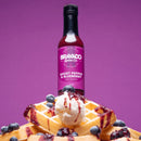 A bottle of Bravado Spice Co Ghost Pepper & Blueberry Hot Sauce from Bravado Spice Co is placed on top of waffles with ice cream, blueberries, and berry sauce against a purple background.