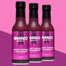 Three bottles of Bravado Spice Co Ghost Pepper & Blueberry Hot Sauce, each featuring vibrant purple labels, are showcased against a striking pink and purple background—the very same bold hot sauce from Bravado Spice Co that took the spotlight on Hot Ones Season 3.