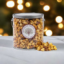 A clear tub labeled Millican Pecan Caramel Pecan Popcorn - 1-Gallon Gift Pail, 1.5 lb sits on marble with caramel popcorn and pecans spilling out, complemented by warm, blurred lights in the background.
