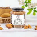 A jar of Millican Pecan Creamy Pecan Butter - 8.5 oz Natural Texas Pecan Spread sits among pecan halves and nuts, with a bowl of pecans and flowering branches softly blurred in the background.