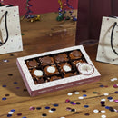 A Millican Pecan Combination 3-Chocolate Caramillicans Gift Box rests on a wooden table sprinkled with confetti, surrounded by colorful gift bags and festive party decorations. The Millican Pecan chocolates are neatly arranged in rows, some adorned with smooth white or rich brown toppings, and feature irresistible pecan candy reminiscent of classic turtle-style confections.