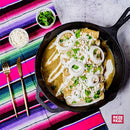 A skillet of enchiladas is topped with onion rings, melted cheese, fresh cilantro, and a savory sauce made using Rico Rico Guajillo Chile Powder Shaker for a mild chili heat and authentic flavor. On the side are small bowls of cheese and cilantro, gold cutlery, and a vibrant striped cloth. The image features the Rico Rico logo—an ideal pick for fans of genuine Mexican cuisine.
