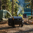 The Green Mountain Grills Trek Prime 2.0 WiFi Portable Wood Pellet BBQ Smoker Grill brings wood-fired flavor to your lakeside campsite, surrounded by pine trees, a green tent, and camp gear—perfect for outdoor grilling adventures.