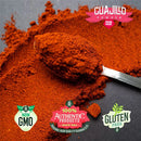A spoon with Rico Rico Guajillo Chile Powder on a dark surface, featuring text that reads "Rico Rico Chile Powder 3 Pack Bundle – Ancho, Guajillo, and Arbol" along with icons for Non-GMO, Authentic Products, and Gluten Free—perfect for adding authentic flavor to your favorite Mexican cuisine.