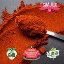 A spoonful of vibrant red guajillo chile powder from the Rico Rico Chile Powder 3 Pack Bundle—featuring Ancho, Guajillo, and Pasilla varieties—is displayed on a dark surface with certifications for Non-GMO, 100% Authentic Products, and Gluten-Free visible nearby. Scattered powder highlights the quality offered by Rico Rico.