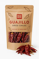 A brown resealable bag labeled Rico Rico Guajillo Dried Chiles – Authentic Medium Heat Mexican Peppers by Rico Rico, with visible dried red chiles inside and several loose guajillo chiles in front—perfect for adding bold, authentic flavor to Mexican dishes with these classic peppers.