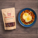 A blue plate with chilaquiles topped with two sunny-side-up eggs sits next to a brown bag of Rico Rico Guajillo Dried Chiles – Authentic Medium Heat Mexican Peppers, emphasizing the essential role of Rico Rico’s peppers in authentic Mexican cooking.