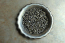 A metal bowl filled with curled, dried World Spice Gunpowder Green Tea leaves sits on a brown textured surface.
