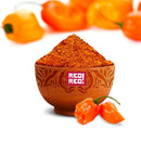 A wooden bowl filled with Rico Rico Habanero Chile Powder sits labeled “Rico Rico”; two fresh orange habanero peppers rest beside the bowl, with more peppers softly blurred in the background. This authentic Rico Rico Habanero Chile Powder Shaker delivers a fiery spice kick that perfectly captures the vibrant flavor of real habaneros.