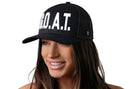 A woman with long brown hair smiles while wearing a black Haka Hat G.O.A.T. Trucker Hat snapback with white lettering. The background is white.