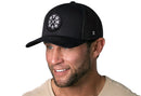 A smiling man with a short beard wears the Haka Hat GYM Trucker Hat, a black workout snapback by Haka Hat with a circular gym-themed logo, paired with a beige shirt against a white background.
