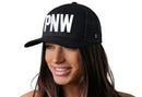 A smiling woman wears the Haka Hat PNW Trucker Hat—also called the Black PNW Snapback—with bold 3D white PNW lettering, shown against a white background.