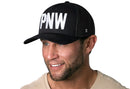 A smiling man wears a Haka Hat PNW Trucker Hat, a black snapback with bold 3D embroidery, and a light shirt against a white background.