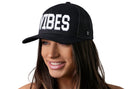A woman with long brown hair wears the Haka Hat VIBES Trucker Hat, a black Good Times snapback featuring "VIBES" in bold white letters. It has a comfort-fit sweatband and is shown against a white background.