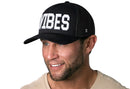 A smiling man with light facial hair wears the Haka Hat VIBES Trucker Hat, a black mesh snapback with white block letters, paired with a light shirt and photographed against a plain white background.