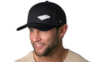 A smiling man wears the Haka Hat Cornhole Board Trucker Hat | Black Bags Snapback by Haka Hat, featuring a white cornhole board graphic on the black mesh front, shown against a white background.