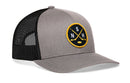 The Haka Hat Nashville Hockey Trucker Hat | NSH X Snapback by Haka Hat features a gray and black design with a round front patch showcasing two crossed hockey sticks, a puck, and the white letters “N,” “S,” and “H” on a yellow border. Part of the Haka signature 5.0 collection, this snapback trucker hat combines classic style with premium comfort.