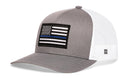The Haka Hat Thin Blue Line Trucker Hat is a gray and white police snapback with an American flag patch featuring a blue stripe. The mesh back offers breathability, making it a perfect tribute to law enforcement.