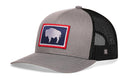 The Haka Hat Wyoming Flag Trucker Hat is a gray and black snapback featuring a mesh back and a front patch with a white buffalo on blue, bordered in red—an iconic nod to the Wyoming flag.