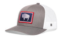 The Haka Hat Wyoming Flag Trucker Hat features a gray-white snapback design with mesh back and a front rectangular patch of a white buffalo on blue, outlined in red.