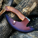 The Texan Knives Custom Hand-Engraved Axe Pizza Cutter, featuring an 8" J2 steel blade, sits on rough tree bark outdoors with its ornate brown leather sheath and curved wooden handle.