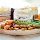 A jar of Terrapin Ridge Farms Hatch Chile Cream Cheese Dip sits on a wooden platter with baby carrots, pretzels, and cucumber sticks. Other Terrapin Ridge Farms Southwest Flavor Dips are blurred in the background.